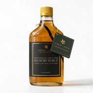 Bottle of Arbor and Field Black Label Bourbon-Infused Shagbark Hickory Syrup with black and gold foil label. A piece if bourbon-infused shagbark hickory bark can be seen inside of the bottle. A green gift tag hangs from the neck of the bottle on a gold string. Clean white background, premium look and feel.