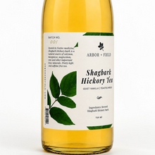Load image into Gallery viewer, Close up of bottle of Arbor and Field shagbark hickory tea with a green and white label and handwritten batch number on the left side. Modern clear glass wine bottle with amber colored tea inside. Clean white background, premium look and feel. 
